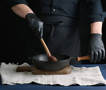 男子用黑色制服和乳胶手套做饭 激起旧木头煎锅烹饪工具勺子乡村食物家庭桌子金属厨具图片