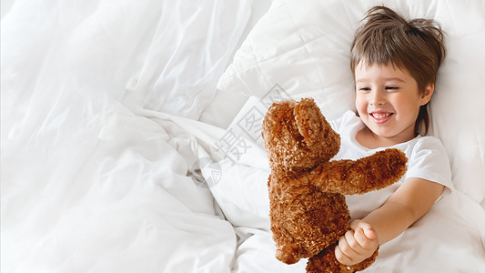 蹒跚学步的孩子和可爱的泰迪熊躺在床上 在白色毯子下的小男孩带着毛茸茸的玩具 毛绒护罩可保护孩子的睡眠 早上在舒适的家里睡觉动物笑图片