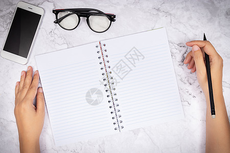 平躺在白色大理石桌子上的空白白页笔记本上的女性手写 顶视图 办公桌上的眼镜和智能手机装饰图片