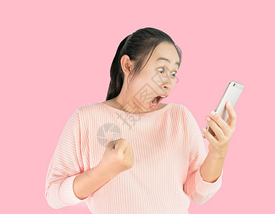亚洲女性震惊了她在智能手机上所看到的 并以粉红色背景孤立地展示了成功的迹象图片