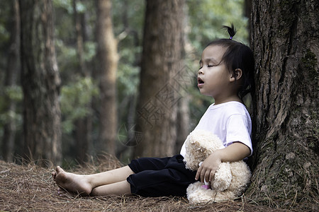 亚洲小女孩正在抱抱熊 独自坐在松树下的地上 笑声学习环境公园家庭情感教育孤独女性女孩木头图片