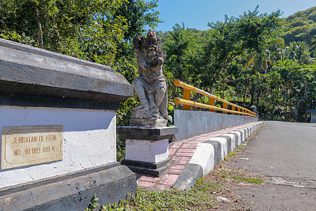巴厘岛山区河流上带黄色金属栏杆的桥 装饰石像代表神话中的保护者 在背景野生热带植被和山脉中雕像文化宗教海岸晴天森林雕塑石头建筑学图片