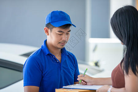 亚洲送货员身穿蓝色制服 头戴蓝帽 手拿纸箱给屋前的女顾客 网上购物和快递房子工作购物服务电子商务女孩送货命令信使船运图片