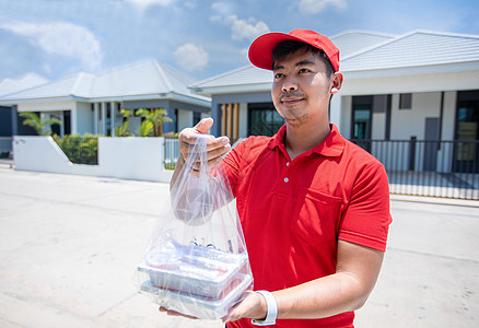 亚洲送货员身穿红色制服 头戴红色帽子 拿着装在塑料袋中的食品盒 交给屋前的顾客 网上购物和快递命令杂货店邮政邮差送货电子商务服务图片