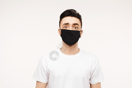 戴着医用面具的害怕的人看着镜头 害怕冠状病毒的害怕的年轻人穿着白色 T 恤和防病毒医用口罩 在白色背景中被隔离 有复制空间图片