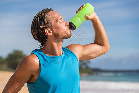 运动瓶饮料运动男子饮用水在海滩上运行 男性跑步者在艰苦的锻炼后出汗和口渴图片