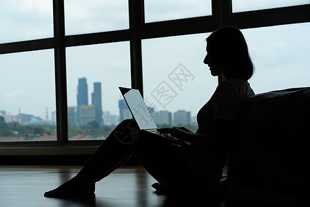 Siluet 女孩在笔记本电脑上工作 喝着咖啡 坐在床边的地板上 靠近全景窗 从高楼层可以看到美丽的景色 时尚的现代内饰女性微笑图片