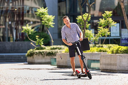 城市中心年轻休闲活跃的运动型商务人士 穿着衬衫和短裤 拿着笔记本电脑包 在炎热的夏日骑着电动滑板车上班图片