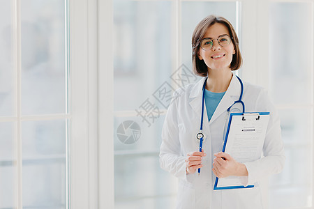 漂亮的微笑实习生拿着写有纸和笔的剪贴板 穿着白色制服 准备在自己的私人诊所看病人 站在室内白色背景下 复制空间区域图片