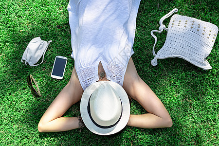 一位白人妇女躺在公园的绿地上休息 脸上戴着白色的帽子 旁边是她的白色巴拿马帽和 COVID 防护面具 — 城市度假安全图片