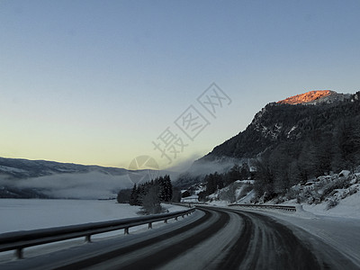 日出时驾车穿过挪威的山脉和森林雪景树木旅行太阳阳光街道运输农村蓝色爬坡图片