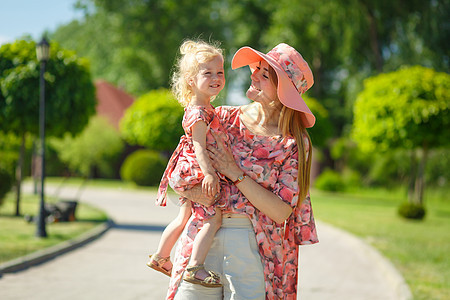 一个穿着浅色夏日太阳裙的迷人女孩和她的小女儿在一个绿色的公园里散步 把她抱在怀里 享受温暖阳光明媚的夏日帽子孩子婴儿童年家庭妈妈图片