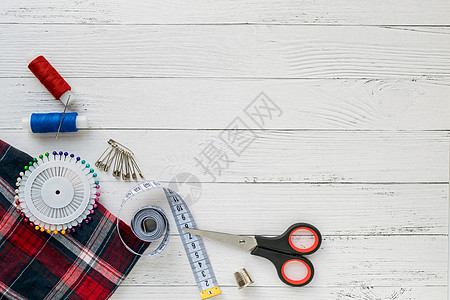 缝纫配件 白色木制背景上的格子织物 织物 缝纫线 针 别针 剪刀和缝纫厘米 顶视图 平面图片
