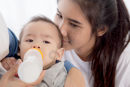 年轻的亚洲母亲在家里用奶瓶拥抱和喂养小女婴 新生儿的纯真与妈妈满意地喝酒 妈妈和孩子的关系和结合 家庭观念童年成人房间女士婴儿投图片