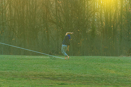 英俊的年轻人在公园里走着滑绳乐趣树干特技赤脚绳索牛仔裤细线钢丝运动成人图片