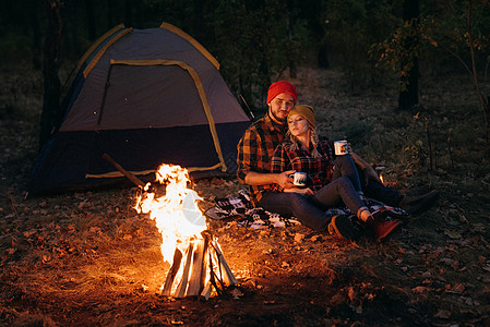 一对年轻夫妇 一个男人和一个女孩 戴着漂亮的编织帽子 在露营时停下来了帐篷女孩篝火金发女郎蓝色旅行胡须橙子取暖温暖图片