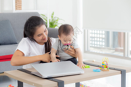 年轻的亚洲母亲在笔记本电脑上工作 同时在家照顾女儿 妈妈和女婴坐在一起使用笔记本视频通话 父母和亲密关系 室内 家庭和商业概念房图片