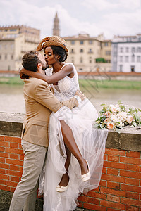 混血新婚夫妇 在意大利佛罗伦萨举行的婚礼 一位非裔美国新娘坐在砖墙上 白人新郎拥抱着她 亚诺河堤岸 俯瞰城市和桥梁图片