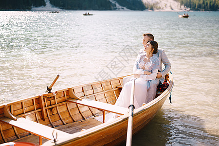 一对新婚夫妇坐在意大利布拉埃斯湖的木船上 新婚夫妇在欧洲 在多洛米蒂山的布雷斯湖上 新郎拥抱新娘恋人婚姻微笑婚礼丈夫假期新人景色图片