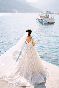 新娘站在黑山佩拉斯特市的码头上 欣赏群山和停泊的船只 裙摆铺在地板上 长长的面纱在风中展开图片