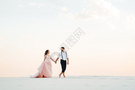 粉色天空一对年轻夫妇 一个穿黑裤子的男人和一个穿粉红色裙子的女孩 沿着白沙走来沙滩卷曲马裤跑步黑色脚印树木胡须青年白色背景