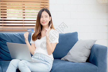 年轻的亚洲女性坐着思考想法 使用信用卡和沙发上的笔记本电脑 女孩在线购物购买和支付 沙发上的笔记本 金融和借记卡 生活方式概念销图片