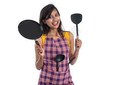 以白色背景持有厨房用的年轻印度妇女SPO STPULA 拖拉机和锅等女孩女性围裙勺子衣服烹饪微笑平底锅肩胛骨餐具图片