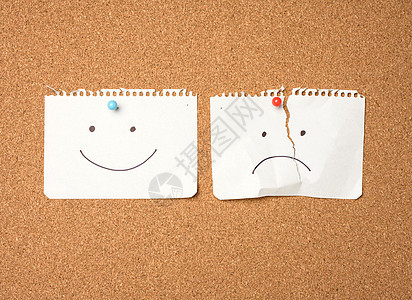 两页纸叶 微笑和悲伤的情绪 与棕色板上的按钮连在一起幸福快乐情感白色棕色软木别针木板图片