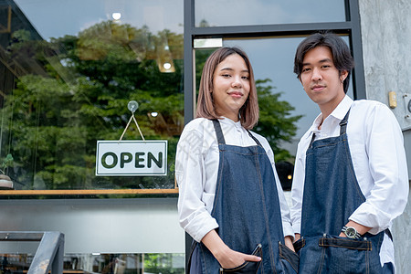 淘宝店铺招牌两名亚洲咖啡师或咖啡制作人以自信行动站立 还透过白天亮的光芒在咖啡店前观看摄影机和微笑背景