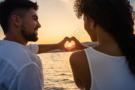 刚结婚的多种族情侣相爱 双手以落日为中心 做心形 看着微笑的眼睛 从后面看西班牙裔女孩和男友的浪漫场景图片