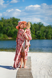 一个穿着浅色夏日太阳裙的迷人女孩和她的小女儿在沙滩上散步 享受温暖阳光明媚的夏日快乐女士海滩母性乐趣裙子婴儿女性拥抱微笑图片