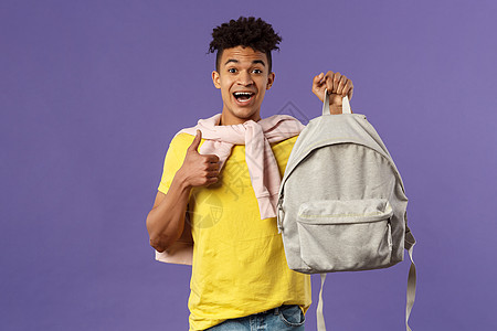 快乐的年轻英俊男学生的画像 男人推荐背包 背着背包 竖起大拇指 为大学学期买了新设备 紫色背景爆炸微笑学习工人发型校园家庭作业培图片