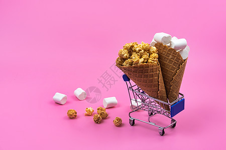 有冰淇淋甜食和粉红背景糖果的购物车零售市场购物粉红色爆米花店铺食物锥体小吃产品图片