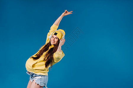 一个穿黄色夹克和耳机的怀孕女孩 站在蓝色背景上女士微笑立体声唱歌帽子休息乐趣旋律音乐衣服图片