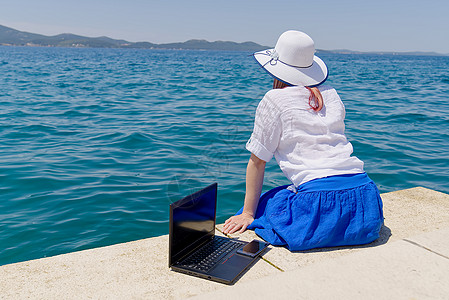 随时随地工作 年轻女性的侧视图 戴着草帽的女性自由职业者坐在沙滩上用笔记本电脑工作 在海边远程使用笔记本电脑工作的年轻女性科技游图片