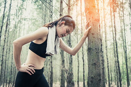 亚洲美女因在森林里慢跑而疲惫不堪 靠在树上 毛巾和汗元素 运动和健康的概念 慢跑和跑步的概念 放松和腰痛主题 户外活动主题图片