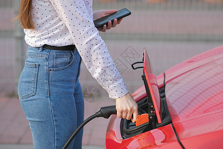 使用智能手机和等待电源连接到电动汽车的白人女孩 汽车充电过程结束图片