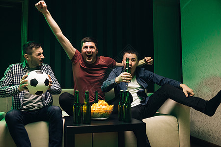 三个英俊 快乐的caucasian友好男子坐在沙发上吃薯片零食 在电视上为足球比赛欢呼 (美国著名橄榄球运动员)图片