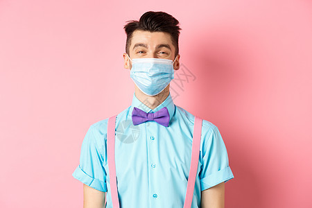 Covid19 大流行病和健康概念 戴医疗面具的微笑者感到快乐 站在节日的蝴蝶领结和吊带上 粉红色背景隔离微笑吊带裤保健工作室广图片