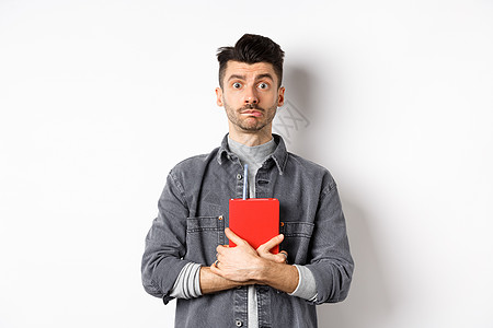 可爱和害羞的家伙拥抱他的日记 拿着红色日记 看相机谦虚 站在白色背景下站立商业男性学生发型办公室胡须日程广告成人胡子背景图片
