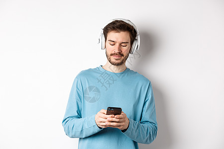 穿着蓝色运动衫的现代青年男子在智能手机上播放音乐 用无线耳机 白背景来听歌曲生活男人成功耳机工作室广告胡须胡子办公室商业背景图片
