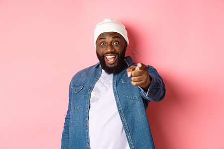 快乐的非洲裔美国潮人用手指指着相机 需要你 微笑着兴奋 站在粉红色背景上男人促销胡须冒充工作室购物横幅帽子衬衫学生背景图片