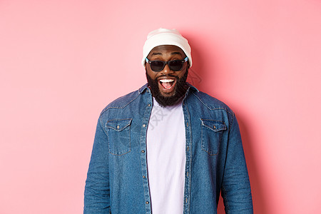 惊讶和快乐的非洲  美国时装男子 在大豆和太阳镜 微笑和看着相机 自信地站在粉红背景上衬衫横幅学生情感潮人成人帽子购物广告男人背景图片