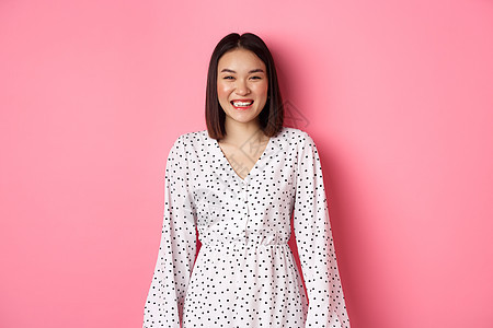 穿着洋装的快乐韩国妇女看着相机 微笑和笑着真诚的表情 站在粉红背景上学生工作室皮肤情感青少年购物广告女孩情绪促销背景图片