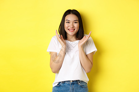 美丽和时尚概念 兴奋的亚洲女性拍手 在相机上微笑快乐 穿着白色T恤站在黄色背景下站立情感黑发理发学生发型女孩交易广告工作室促销图片
