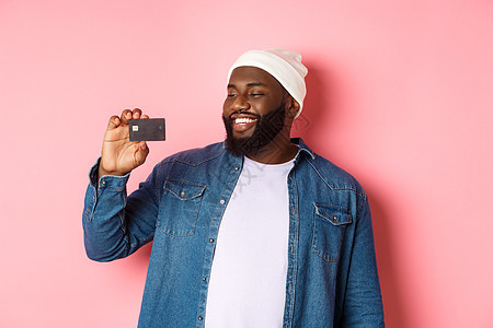购物概念 快乐的非洲美国人对信用卡满意 推荐银行 站在粉红背景上的人的形象 他们向银行提出建议成人促销衬衫帽子情感胡须学生男人横图片