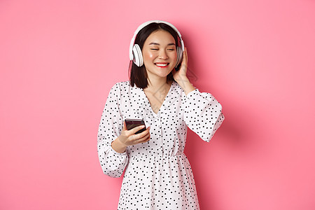 用耳机听音乐 闭着眼睛微笑 握着手机 站在粉红背景上站立的浪漫的亚洲女孩工作室技术快乐购物促销互联网网络电话皮肤女性背景图片