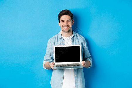 英俊的年轻人在笔记本电脑屏幕上介绍产品 展示计算机和微笑 站在蓝色背景之上情绪成人自由职业者广告手势男性网络商业技术成功图片