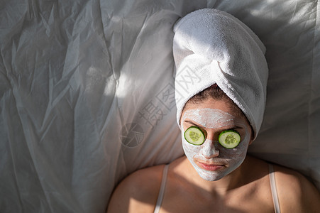 头发有毛巾和粘土中蒙面面具的妇女 当她眼前的黄瓜就躺在床单上奶油惊喜谎言剥皮化妆品呵护黏土卫生护理保湿图片