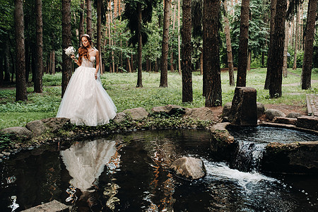 一位身着白色连衣裙 戴着手套 手捧花束的优雅新娘站在森林的溪流旁 享受着大自然 自然公园里穿着婚纱和手套的模特 白俄罗斯赤脚若虫图片